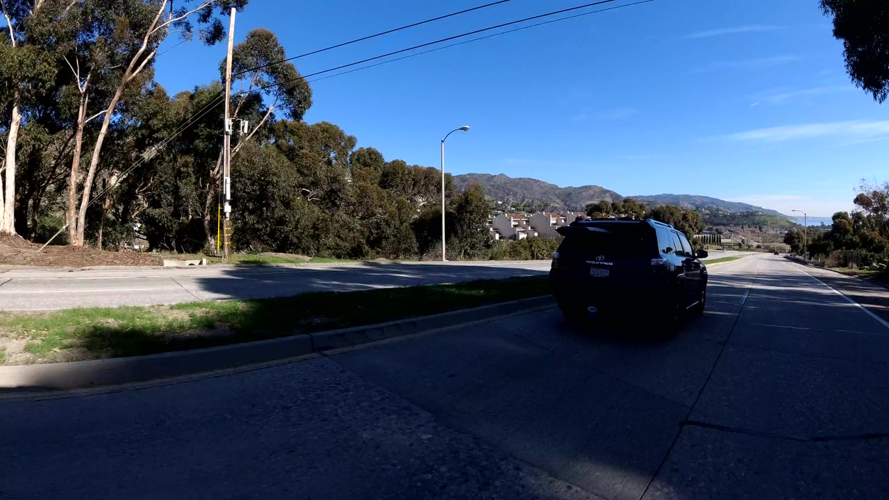 A dark SUV travels along a Malibu roadway with roadside trees, utility poles, overhead wires, distant hills, and deep foreground shadow, with no visible people.