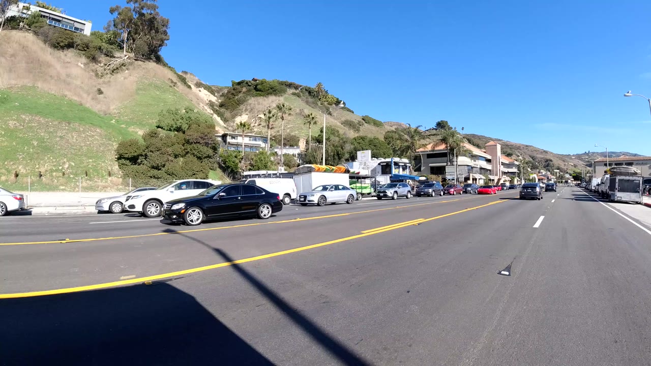 Street-level view of a Malibu road with double yellow lines, parked cars, hillside homes, dry grass slopes, and no visible people.