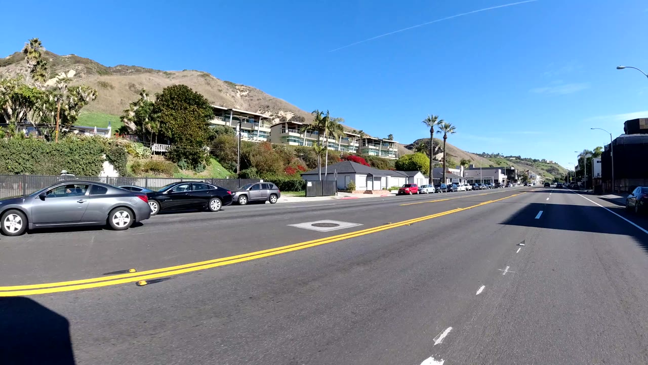 Malibu roadway with a gentle bend, parked cars along the curb, homes at the base of dry hills, palm trees, and no visible people.