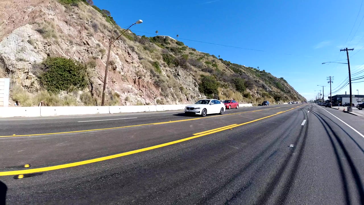 Wide road-level frame in Malibu showing oncoming cars, a broad asphalt lane, yellow center lines, rocky hillsides, power poles, and no visible people.