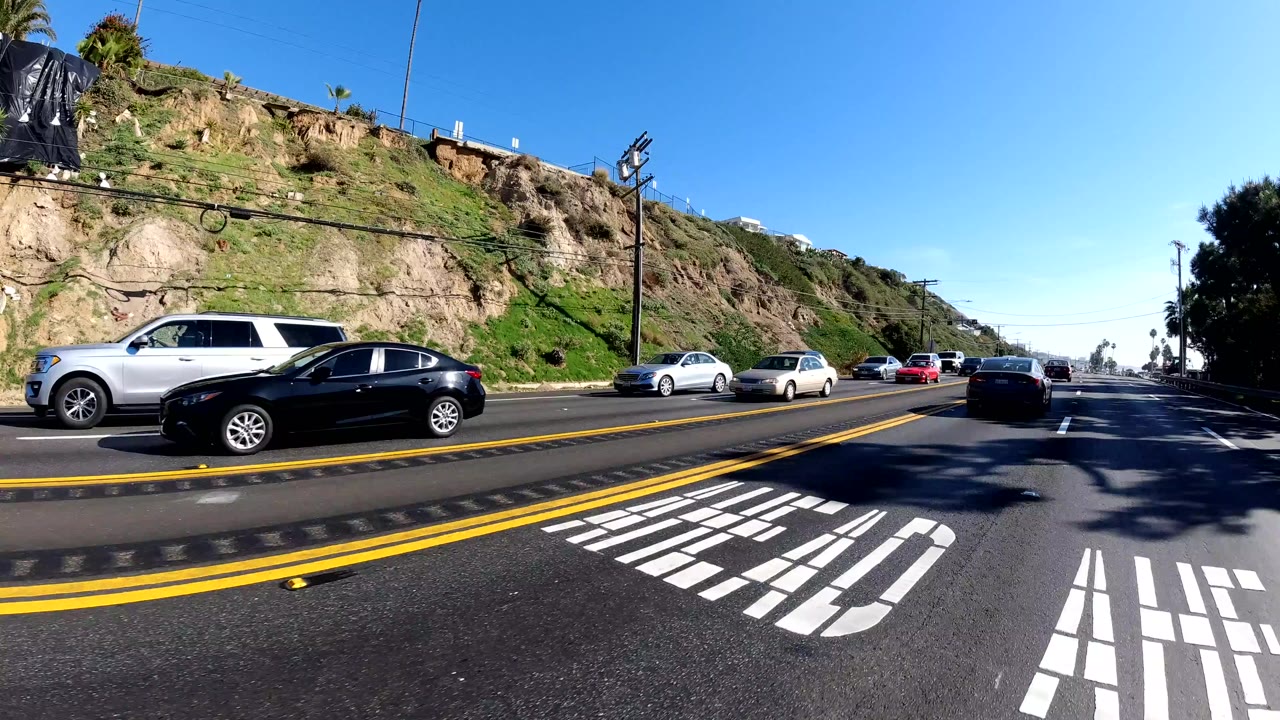 Traffic moves along a multilane road in Malibu, California, with yellow center lines, white road markings, hillside scrub, and no visible people.