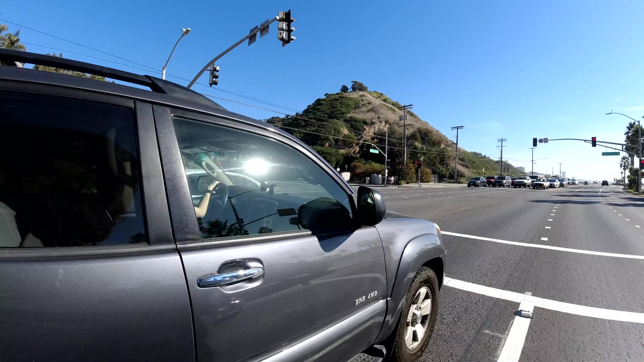 A gray SUV fills the foreground at a Malibu intersection, with traffic lights, lane lines, hillside terrain, and no visible pedestrians.