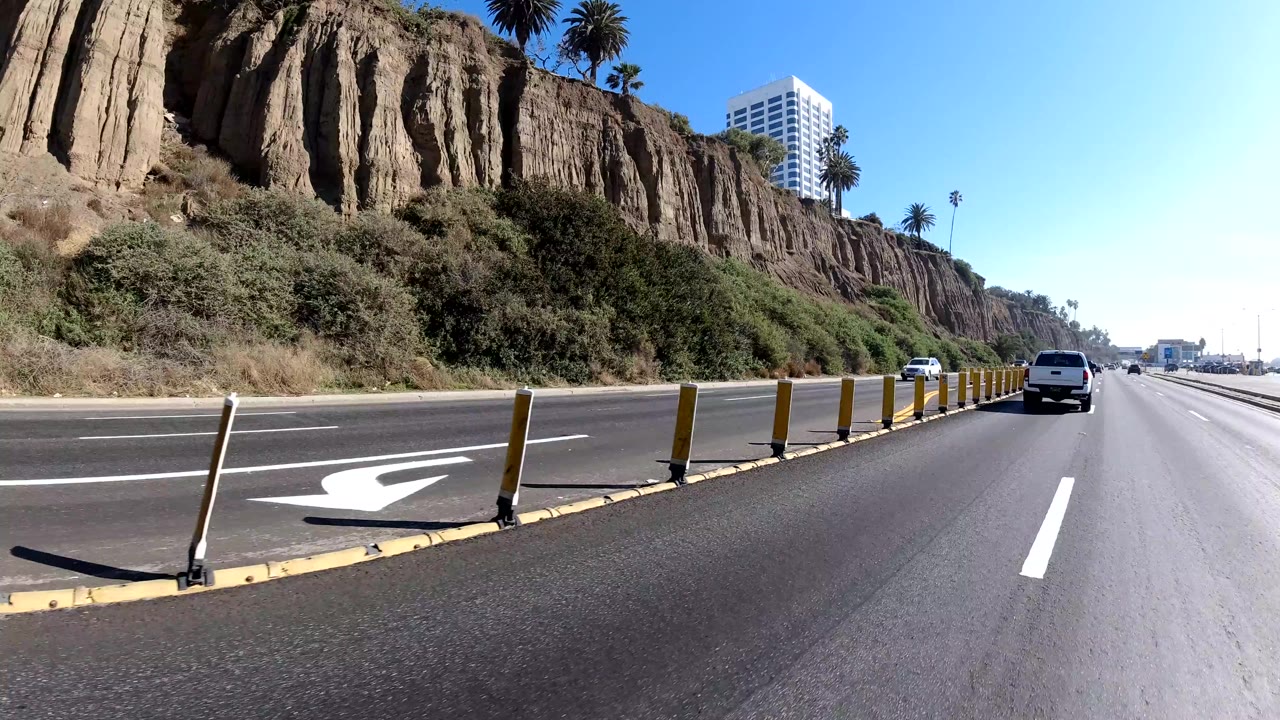 Forward roadway view in Malibu, California showing lane dividers, flexible bollards, dry cliff faces, a white vehicle ahead, palms on the ridge, bright blue sky, and no visible people.