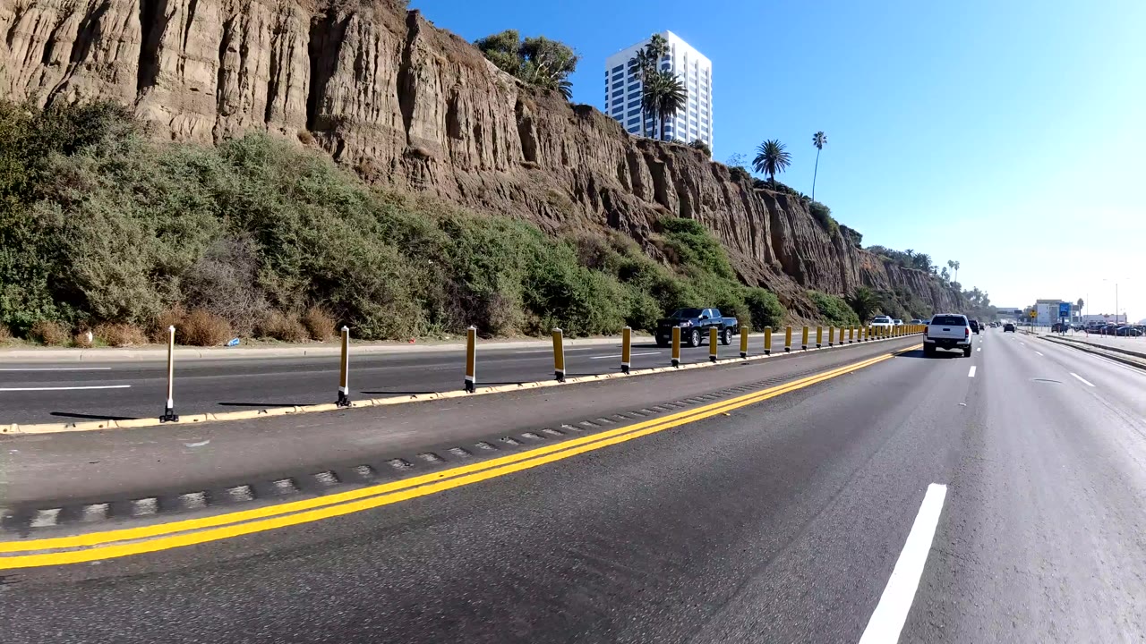 Highway lane curving past bluffs with a white car ahead and a high-rise visible above the ridge.