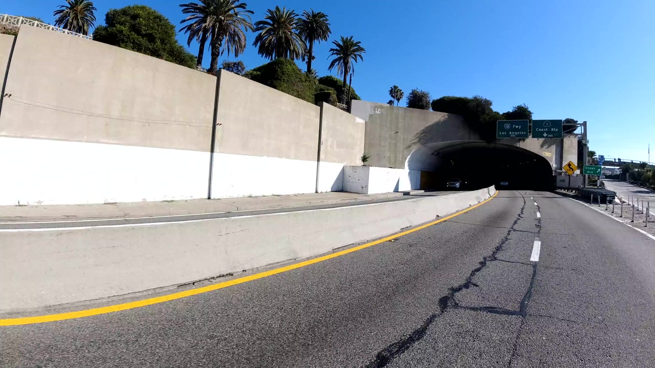 Malibu, California highway scene with a concrete retaining wall, tunnel entrance, curving lane, overhead green freeway signs, palms above the wall, vehicles near the tunnel, and no visible people.
