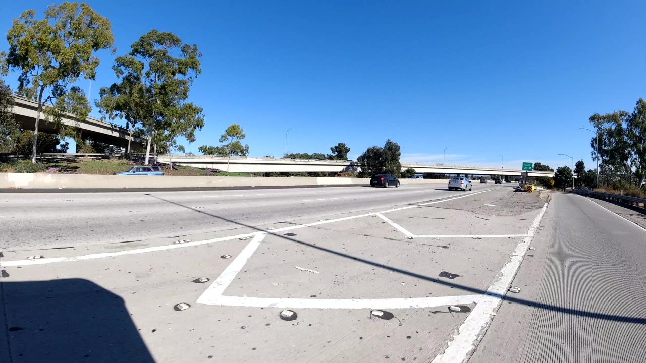 Wide road-level view of a Malibu, California freeway with lane markings, concrete barriers, trees, passing cars, an overpass, and no visible people.