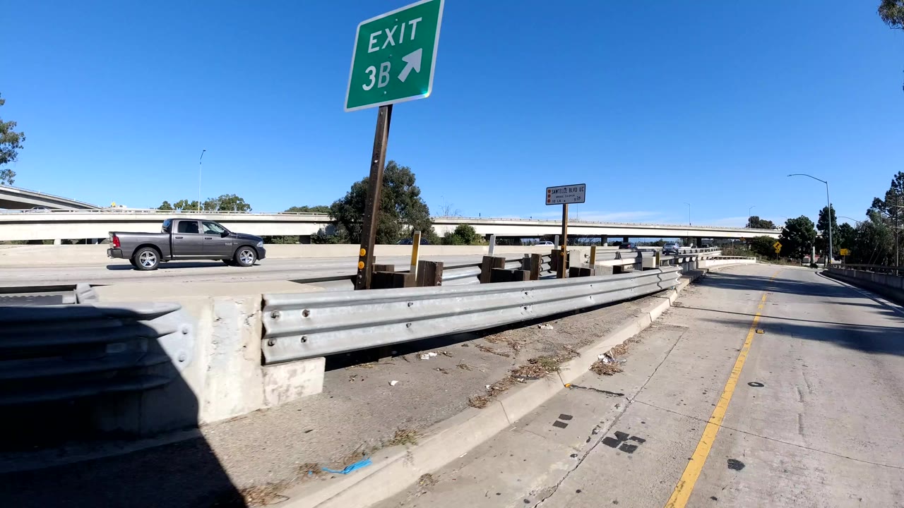 Roadside freeway ramp view in Malibu, California showing a metal guardrail, a green Exit 3B sign, a pickup truck on the elevated road, concrete barriers, and no visible people.