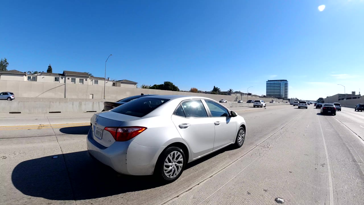 Malibu, California freeway view with a silver sedan close to camera, concrete wall, distant traffic, roadside building, bright sky, and no visible people.