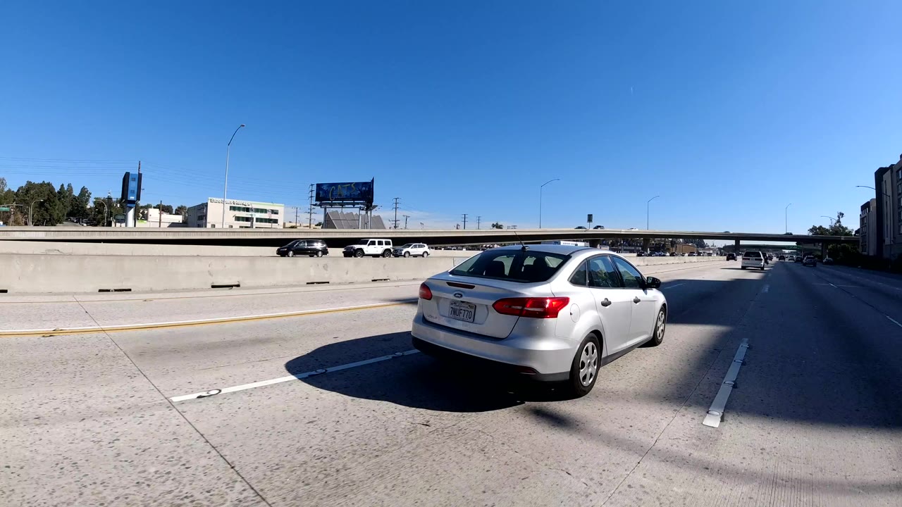 Wide road-level view of cars on a Malibu, California freeway with a silver sedan near the foreground, overpass ahead, blue sky above, and no visible people.