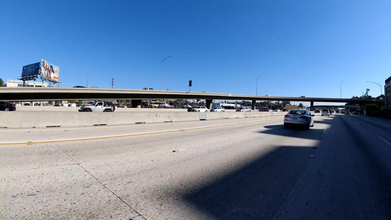 Malibu freeway traffic approaches and passes beneath a concrete overpass, with cars spread across several lanes, billboard structures in the background, and no visible people.