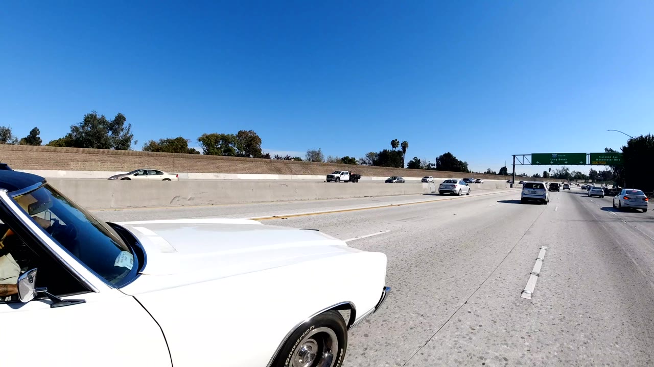 A white classic car fills the right foreground on a Malibu freeway while other vehicles continue ahead across open lanes under a clear blue sky, with no visible people.