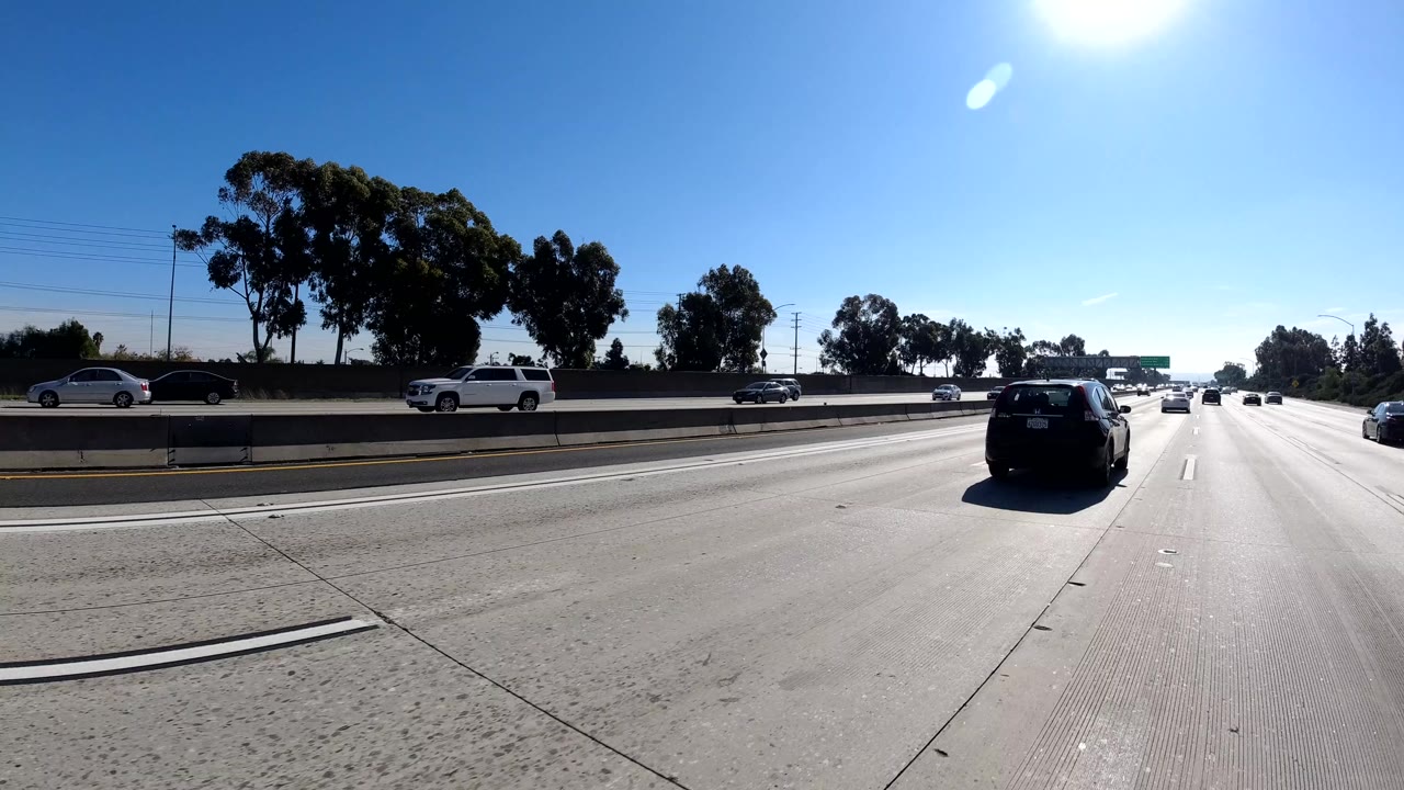 Wide freeway view in Malibu, California with cars ahead and beside the camera, concrete barriers, trees along the side, strong sun glare, and no visible people.