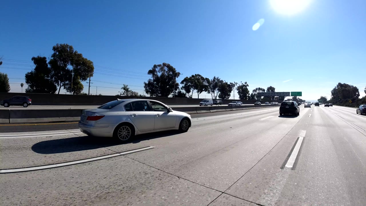 Malibu, California freeway frame with a white sedan close in the adjacent lane, distant cars ahead, tree line beyond the barrier, bright sky, and no visible people.
