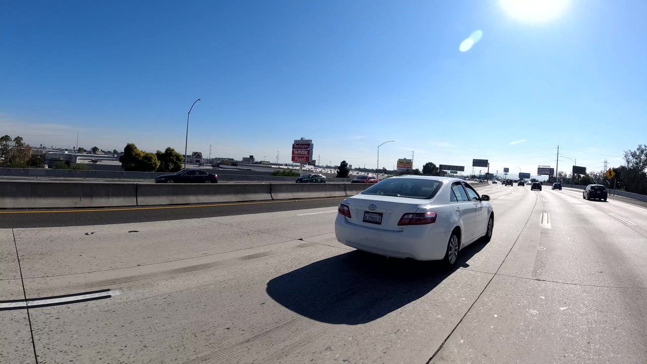 Forward-facing freeway view in Malibu, California with a dark car ahead, overhead road signs in the distance, low concrete dividers, tree silhouettes, sun glare, and no visible people.