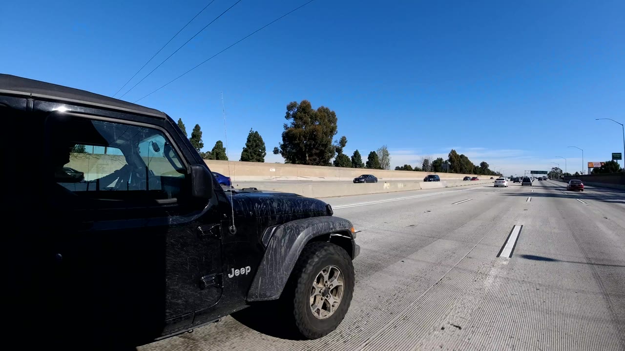 A black Jeep passes close to the camera on a Malibu, California freeway, with open lanes ahead, roadside trees, clear sky, and no visible people.