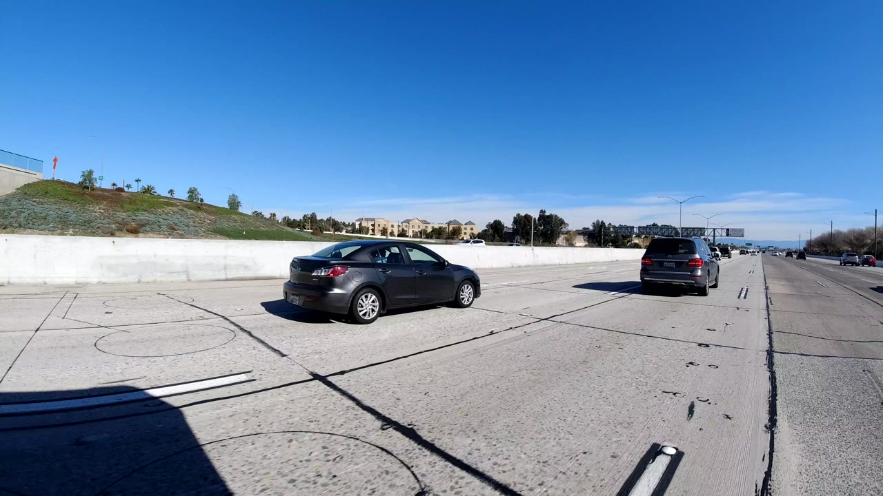 A Malibu, California freeway view shows two nearby cars, concrete lane barriers, hillside buildings in the distance, and no visible people.