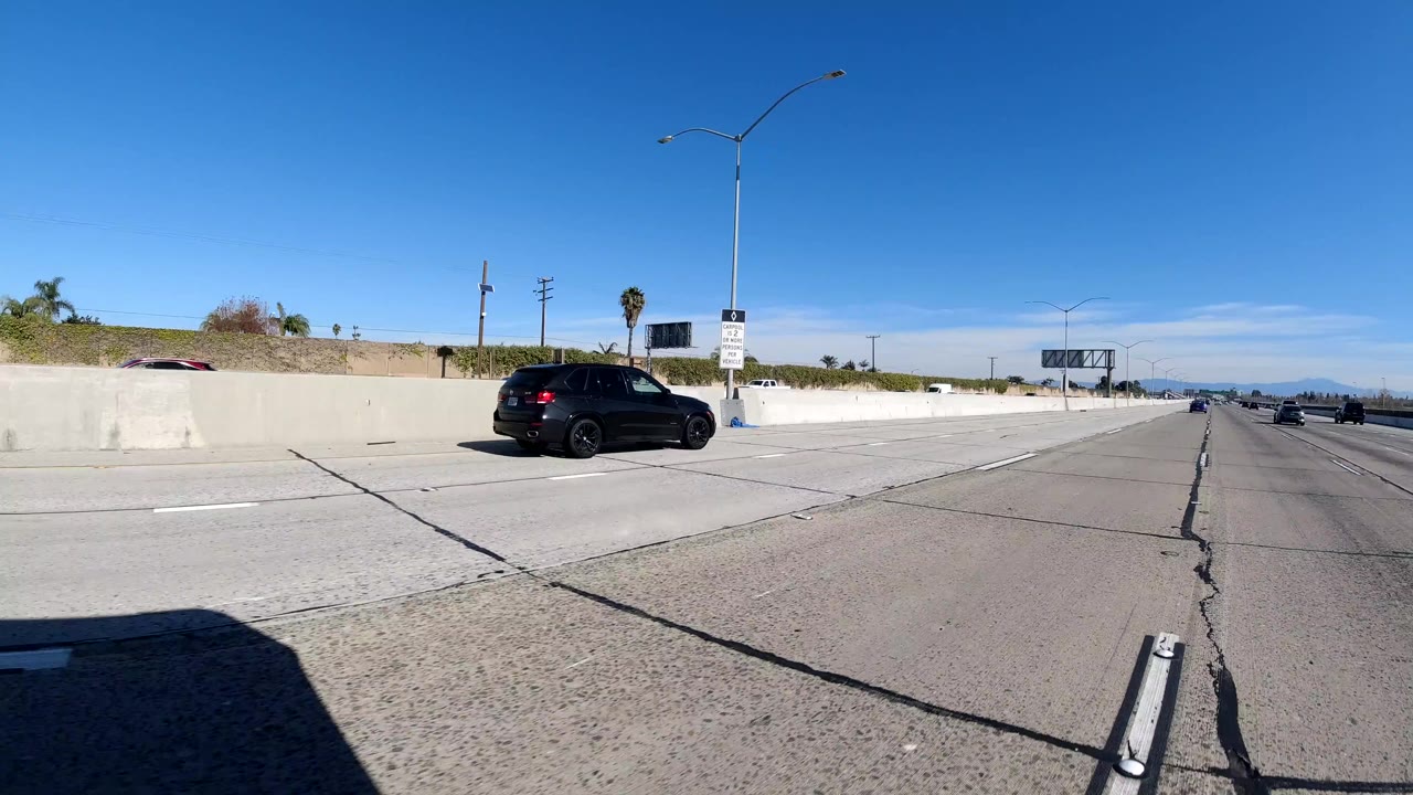 A black SUV passes along a Malibu freeway beside a pale concrete barrier, with roadside signs, light poles, distant traffic, and no visible people.