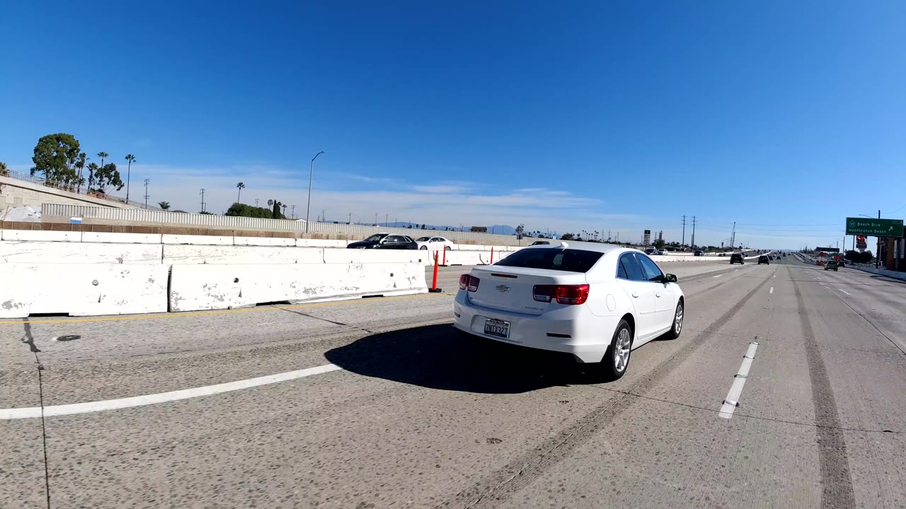 A white sedan passes close on a Malibu, California freeway under a clear blue sky, with concrete dividers, lane markings, distant cars, and no visible people.