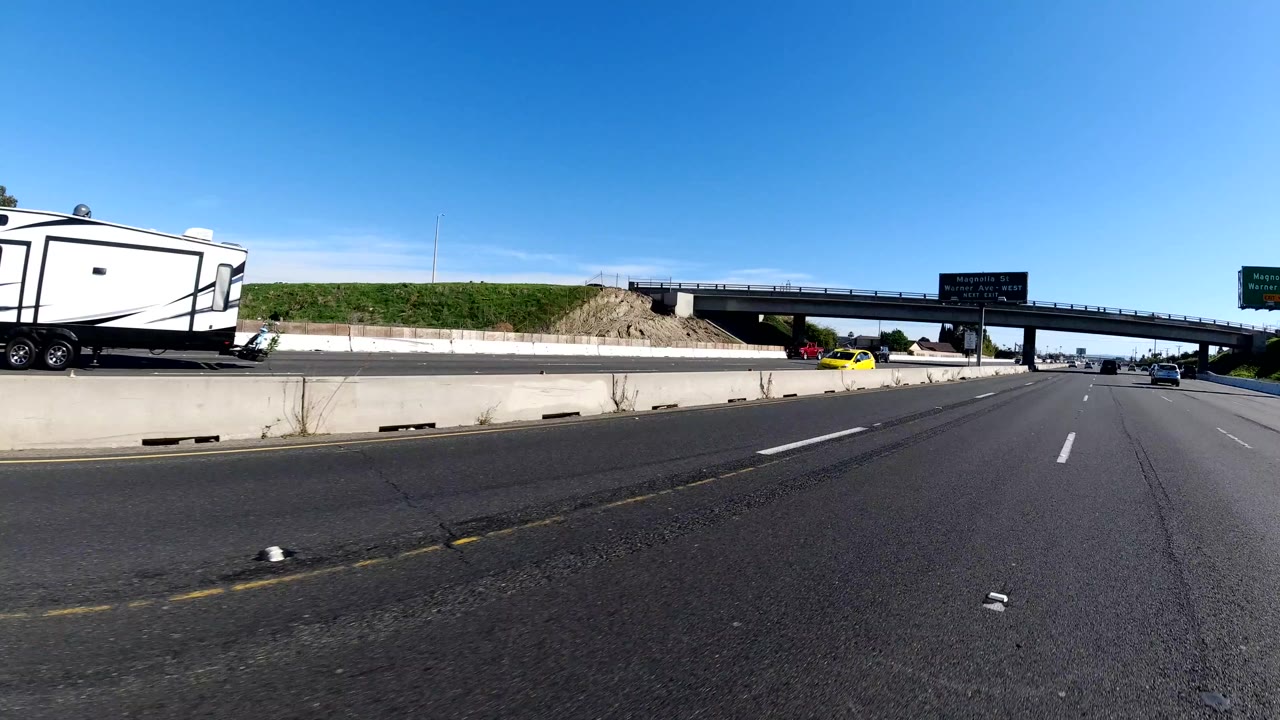 An RV trailer, cars, concrete barriers, an overpass, and freeway signs appear on a bright Malibu highway segment, with no visible people in the frame.
