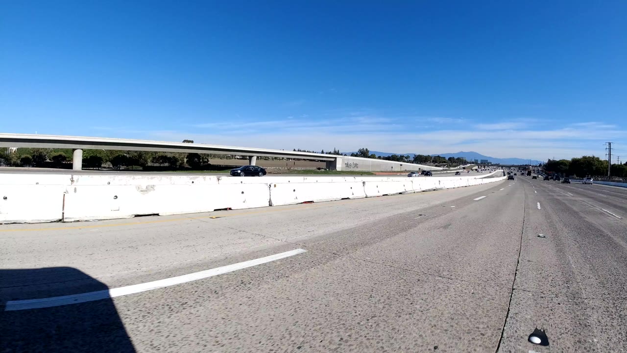 Wide roadway-level view of Malibu freeway lanes with concrete barriers, a distant overpass, scattered cars, bright blue sky, and no visible people.