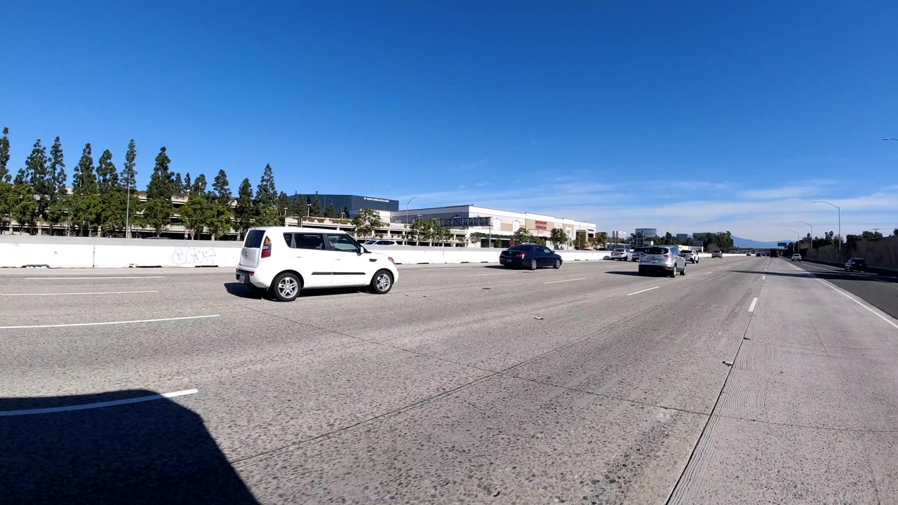 Malibu freeway view with a white van, several cars, roadside trees, modern office buildings, concrete divider, bright sky, and no visible people.