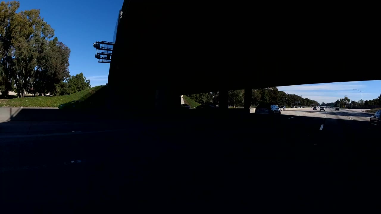 Roadway emerging from heavy bridge shadow with trees and freeway lanes ahead.