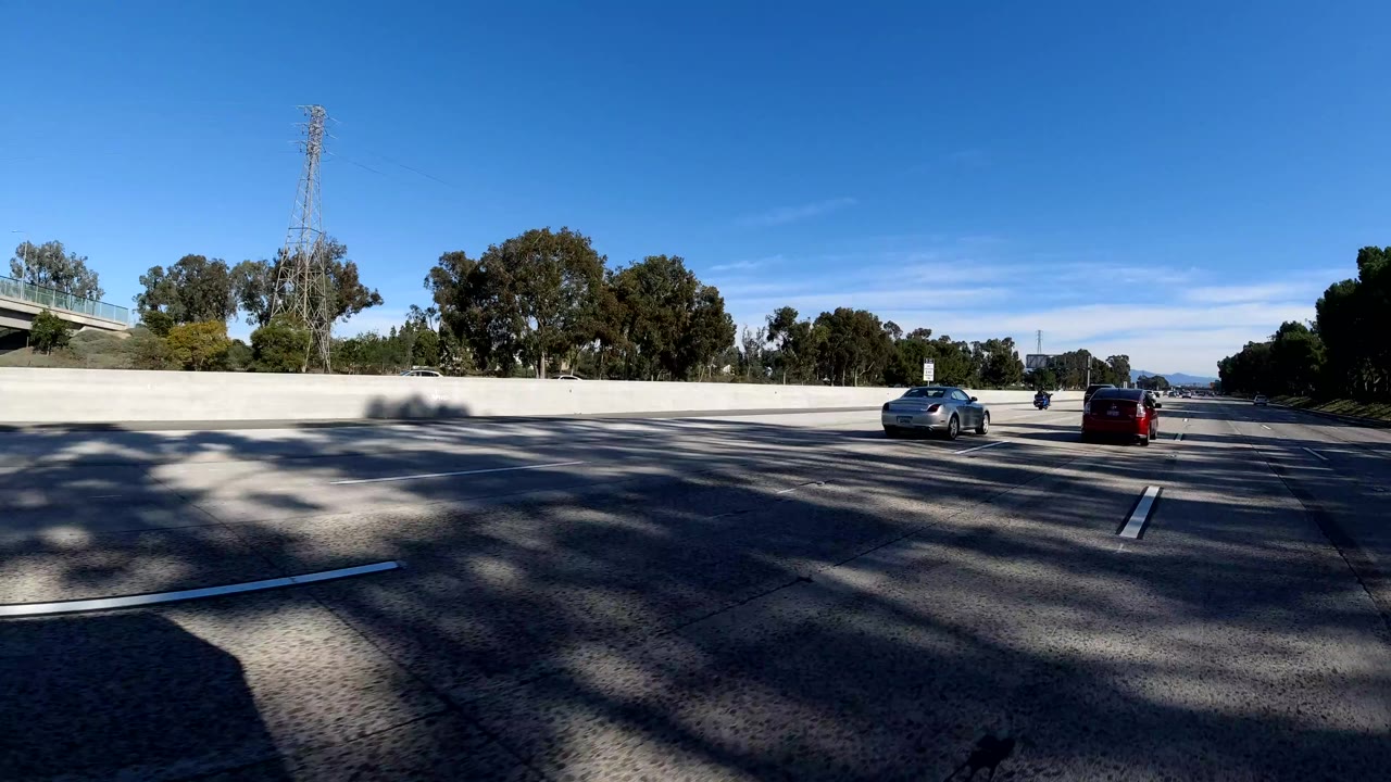 Bright Malibu highway scene with two cars ahead, a concrete median barrier, roadside trees, a power tower, blue sky, and no visible people.