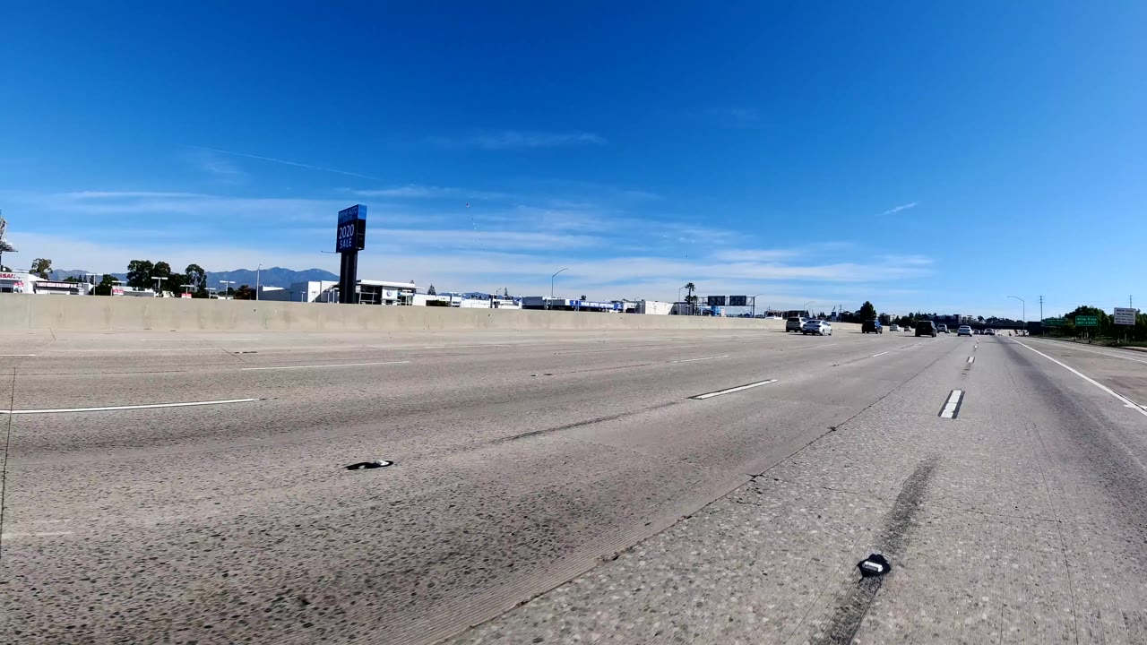 Malibu freeway scene with mostly open lanes, a concrete divider across the midground, distant cars, roadside business signage, mountains faintly visible, and no visible people.