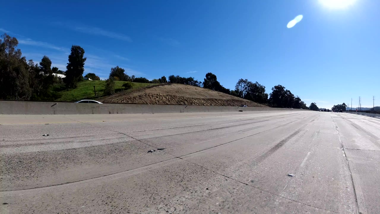 Wide Malibu freeway view with empty foreground lanes, a concrete barrier, green hillside, trees, clear blue sky, and no clearly visible people.