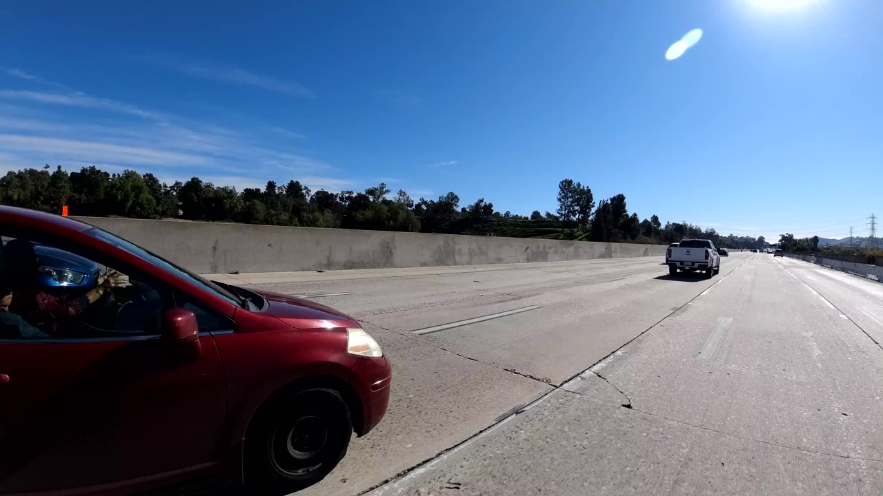 Malibu freeway traffic frame with a red car close at left, a white van ahead, concrete barrier, tree line, bright sun, and no clearly identifiable people.