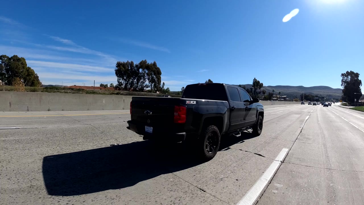 A black pickup truck passes close in the right side of a Malibu freeway frame, with sun glare, concrete barriers, distant hills, and no visible people.