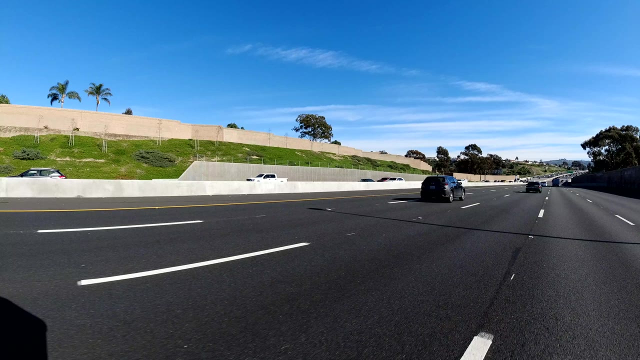 Forward road view on a multi-lane highway in Malibu, California, with cars ahead, a grassy embankment to the left, bright blue sky, and no visible people.