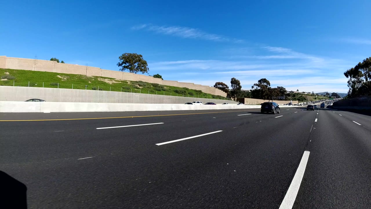 Malibu highway view with dark asphalt lanes, several vehicles ahead, a pale retaining wall beside a green slope, distant trees, and no visible people.