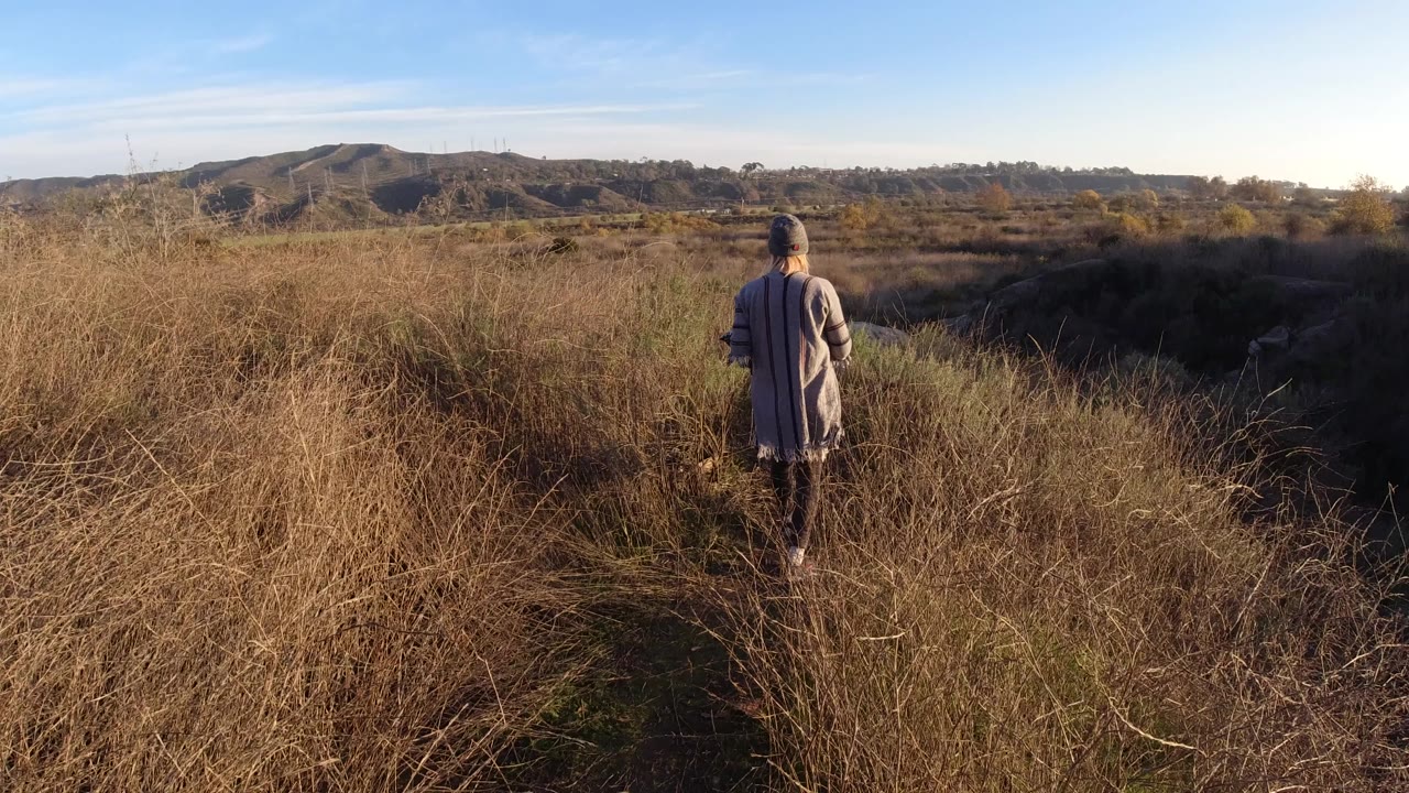 A woman walks away from camera on a dirt path through tall dry grass in San Clemente, California, with low hills and blue sky beyond.
