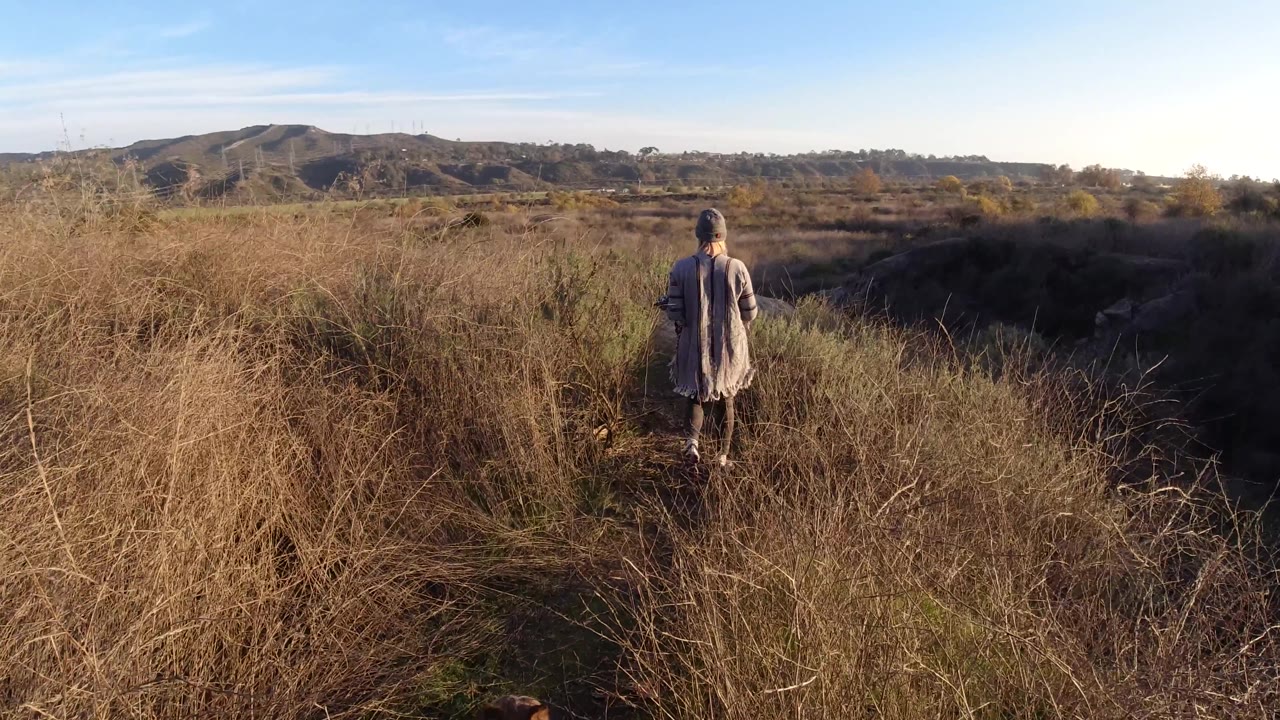 A woman walks near the right side of the frame on a narrow path through dry grass, with a darker ravine area and San Clemente hills behind her.