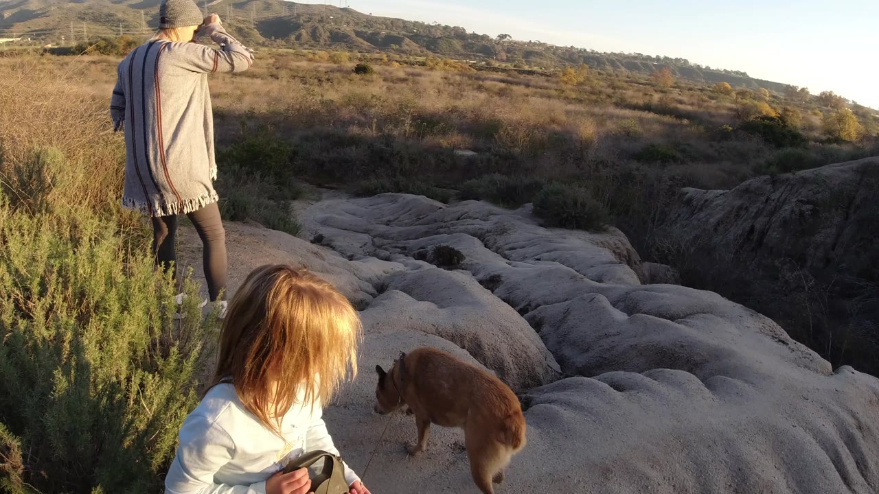 A woman in a knit cap stands ahead of a young girl and brown dog on pale eroded rock beside dry brush in San Clemente, California.