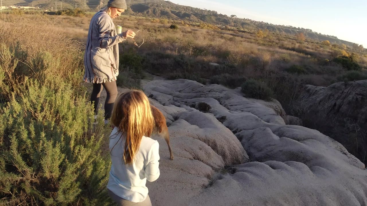 A woman holding small objects stands near a young girl on the ridge above pale rock and dry brush in San Clemente, California.