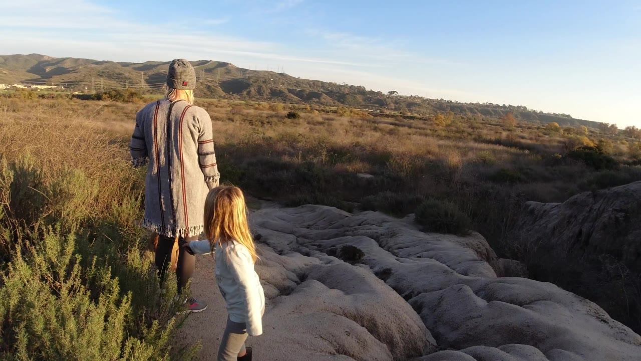 At 0:00, a woman and young girl stand with their backs to camera on pale rock and brush in San Clemente, California, looking toward dry grassland and low hills.