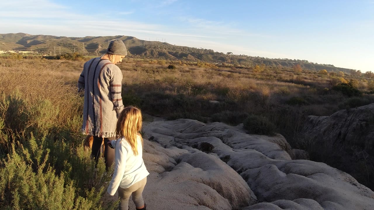 At 0:02, the woman appears turned slightly to her right while the child stands beside the pale rock, with warm sun on their clothing and the San Clemente hills beyond.