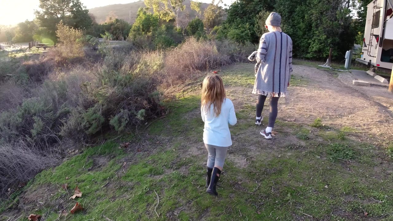 Girl and adult walk away near a parked RV and brush.