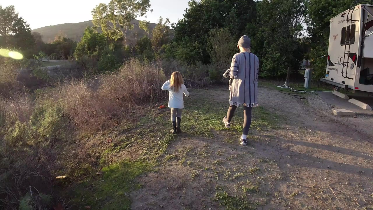 A woman and young girl walk away from camera in San Clemente, California, with dry brush on the left, a dirt path ahead, and a camper at the right edge.