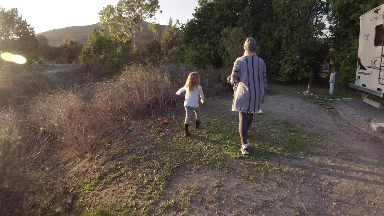 In San Clemente, California, the woman and child are farther along the dirt path, with tall brush at center, trees behind them, utility posts on the right, and the camper partly visible.