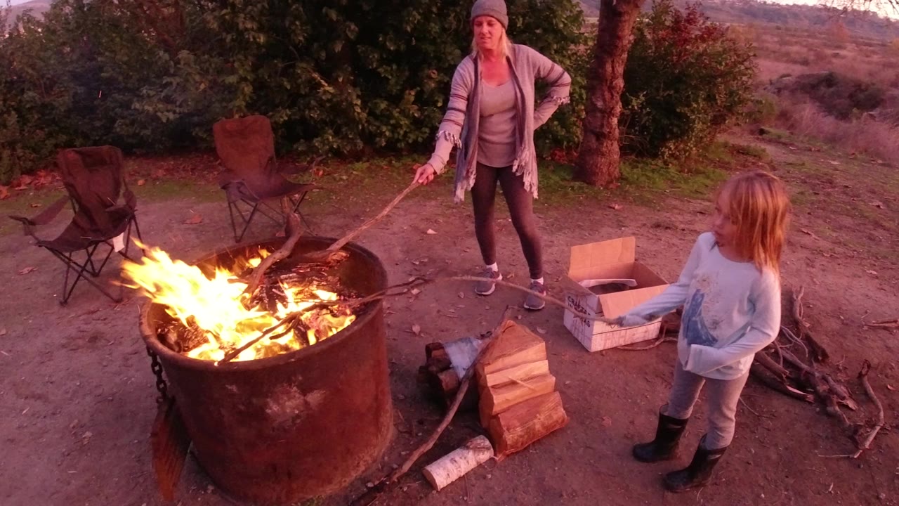 In San Clemente, California, a woman holds a roasting stick beside a large metal fire pit while a child stands at the right edge near stacked firewood and camp chairs.