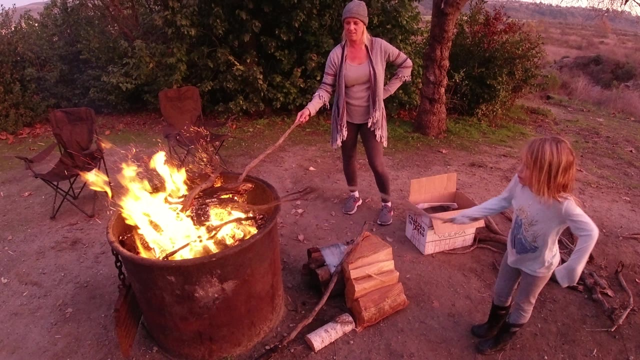 The child reaches a roasting stick toward taller flames while the woman stands behind the fire pit, with chairs, firewood, a box, trees, and hillside visible in San Clemente.