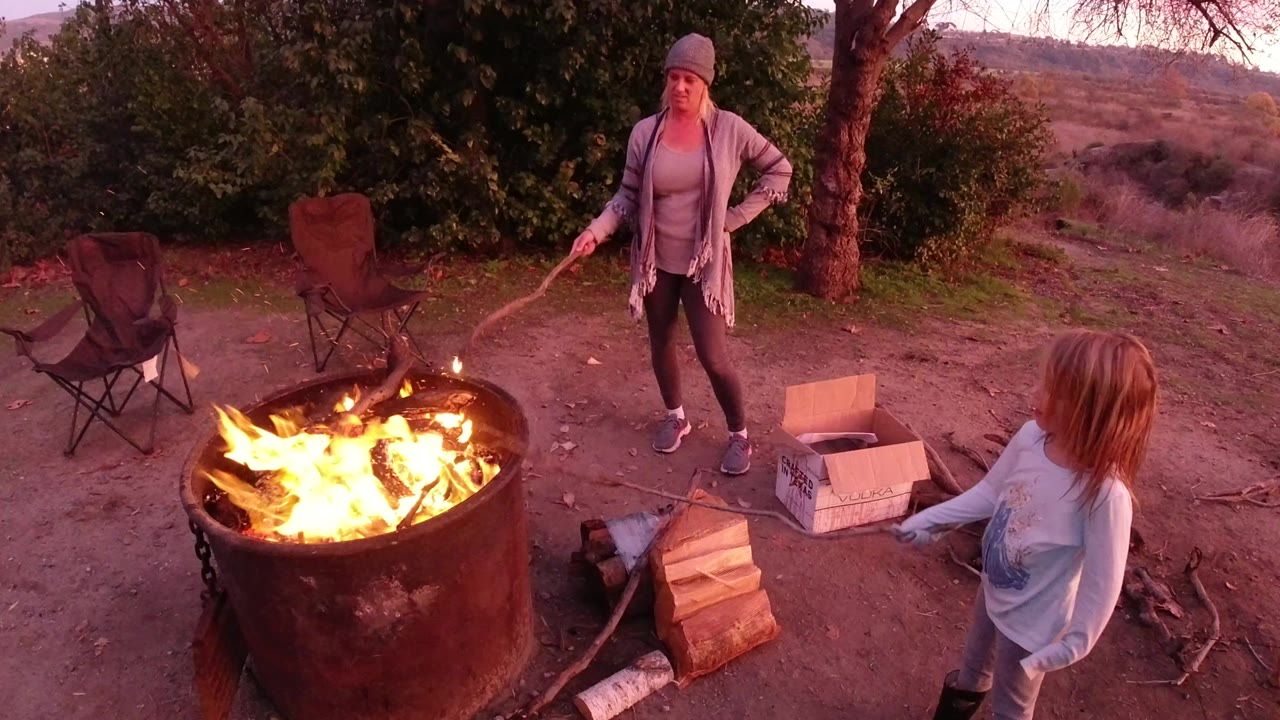 In San Clemente, California, an adult woman stands near a large metal fire pit while a child-height person stands at the right edge beside firewood and cardboard boxes.