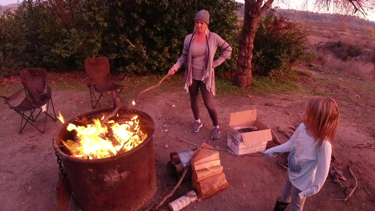 Woman holds a stick near a burning fire pit as another person stands nearby.