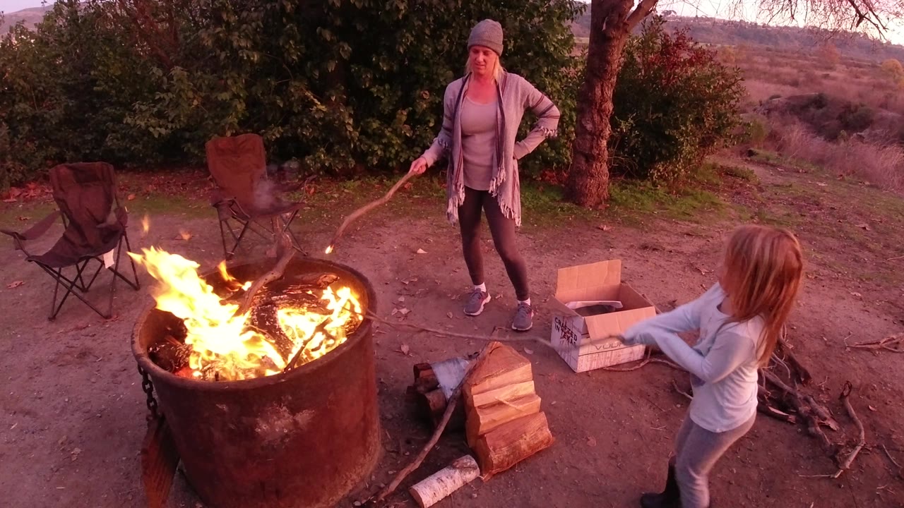 Bright flames flare higher in the metal fire pit as the woman stands behind it and the child-height person remains on the right side of the outdoor San Clemente scene.