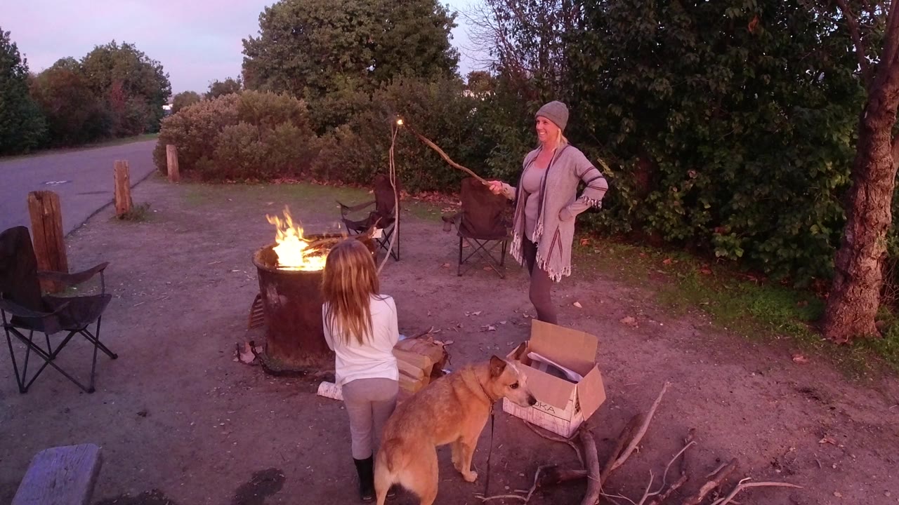 In San Clemente, California, the child remains close to the barrel fire as the woman stands to the right with a long roasting stick and the dog waits near the cardboard box.