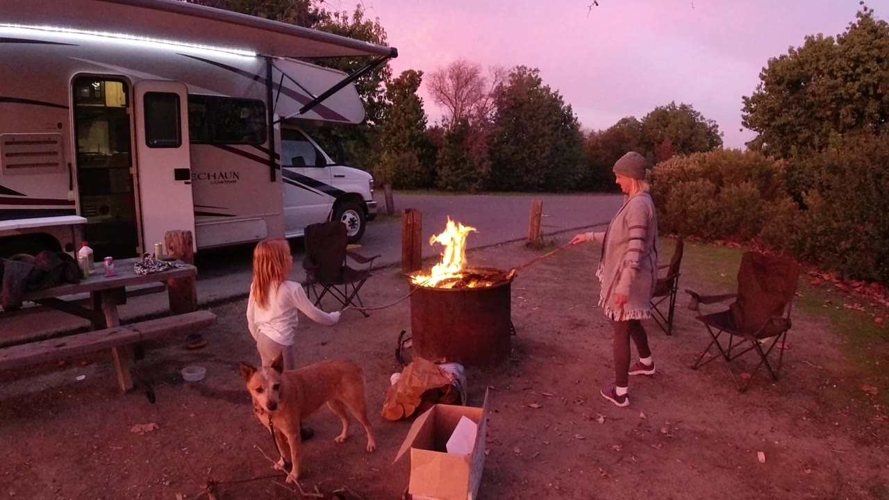 In San Clemente, California, an adult stands near a metal fire barrel while a child sits beside it, with a dog in the foreground and an RV under an awning at dusk.
