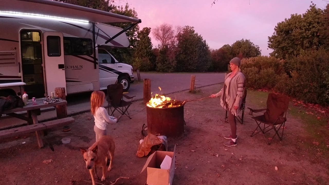 Young girl and dog in foreground with woman standing near a fire pit and RV.
